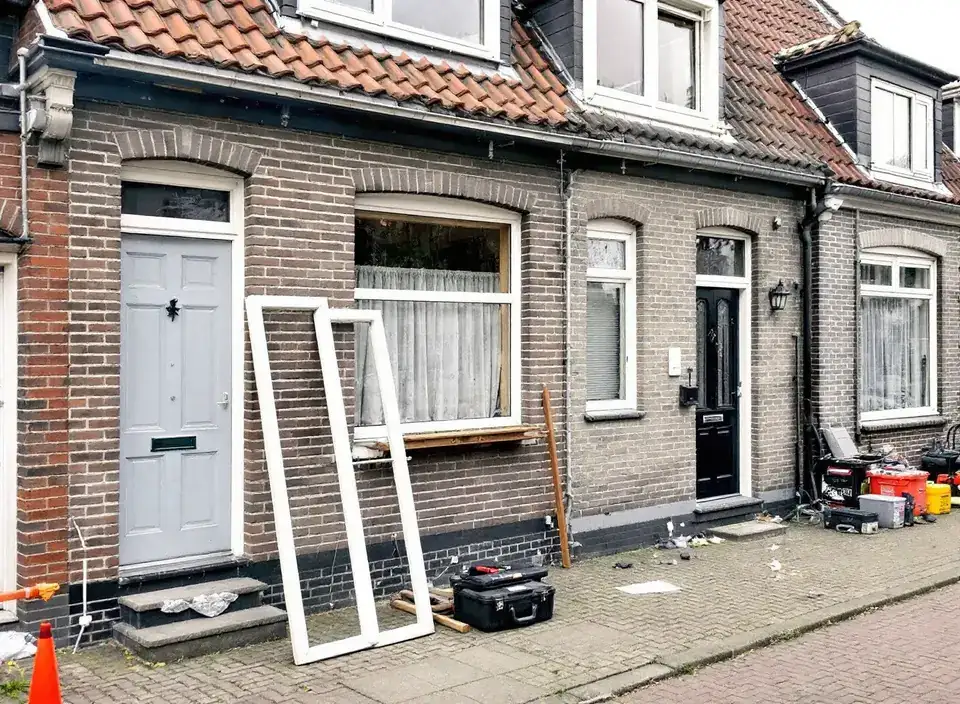 Een rij bakstenen arbeiderswoningen waar de kozijnen worden vervangen: een oud kozijn staat tegen de gevel, gereedschapskisten op de stoep van klinkers
