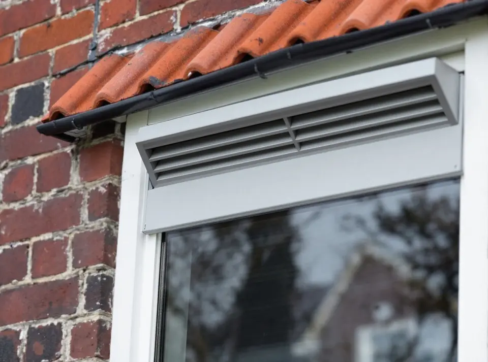 Een raam van een bakstenen rijtjeshuis van buiten, met bovenin het witte kozijn een grijs ventilatierooster met open lamellen onder een smalle regenkap, en oranje dakpannen erboven