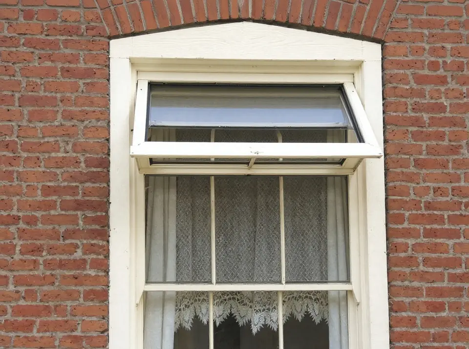 Een klepraam boven een groter raam in een oude rode bakstenen gevel, opengezet, met vitrage erachter
